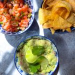 three bowls of guacamole, salsa and chips on a blue counter
